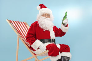 Santa Claus holding a bottle of beer seated on a sun lounger and looking at the camera with blue sky in the background